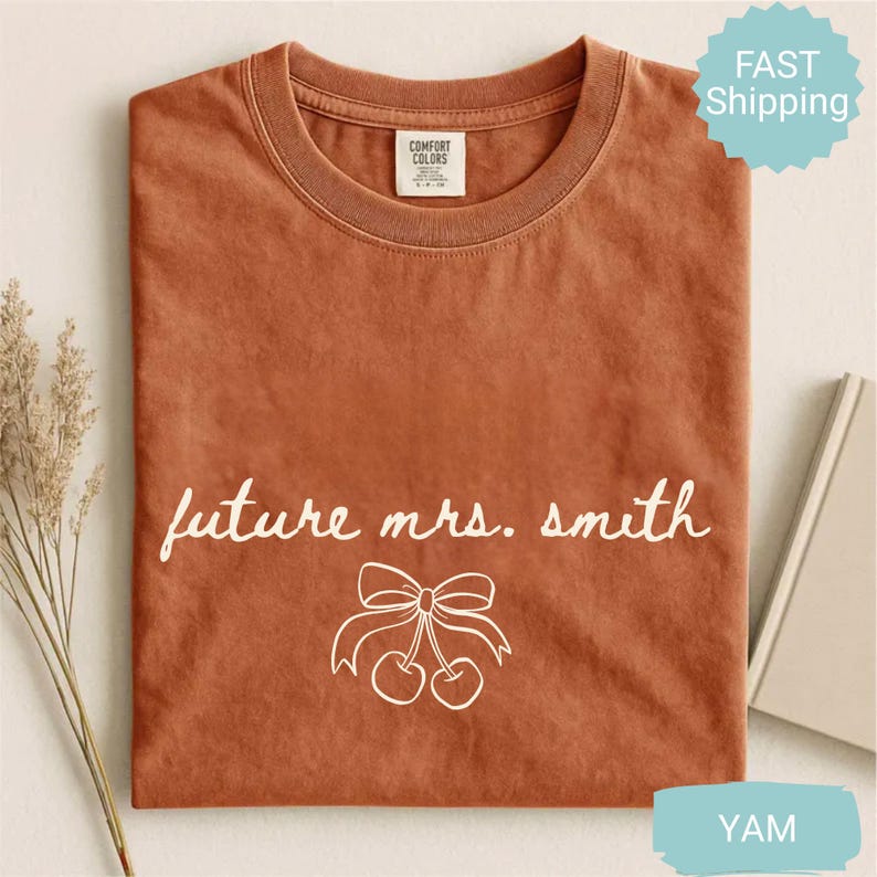 May include: A burnt orange t-shirt with the words "future mrs. smith" in white script. Below the text is a white illustration of cherries and a bow. The shirt has a crew neck and short sleeves. The tag reads "Comfort Colors".