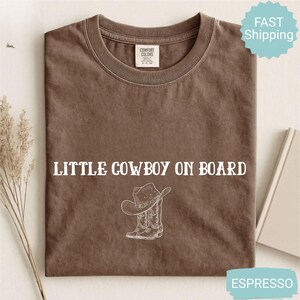 May include: Espresso-colored t-shirt with the text "LITTLE COWBOY ON BOARD" and a graphic of a cowboy hat and boots. The shirt is made of a soft material and is likely pre-shrunk. The shirt is a Comfort Colors brand.