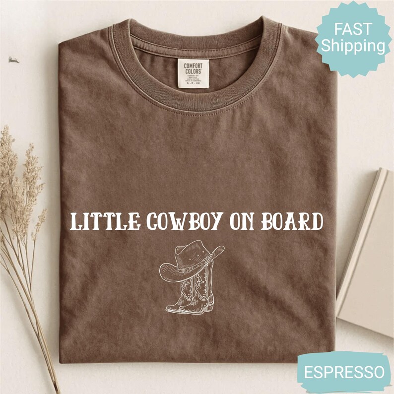 May include: Espresso-colored t-shirt with the text "LITTLE COWBOY ON BOARD" and a graphic of a cowboy hat and boots. The shirt is made of a soft material and is likely pre-shrunk. The shirt is a Comfort Colors brand.
