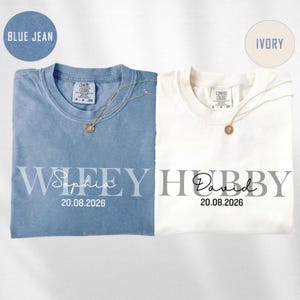 May include: Two t-shirts, one blue and one ivory, with the words "Wifey" and "Hubby" printed on them. The blue shirt has the name "Sophia" and the date "20.08.2026" printed below "Wifey". The ivory shirt has the name "David" and the date "20.08.2026" printed below "Hubby".