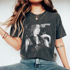 Puede incluir: Camiseta gris oscuro con una imagen en blanco y negro de una persona. La persona lleva gafas y tiene el pelo largo. La camiseta es de estilo vintage con cuello redondo.