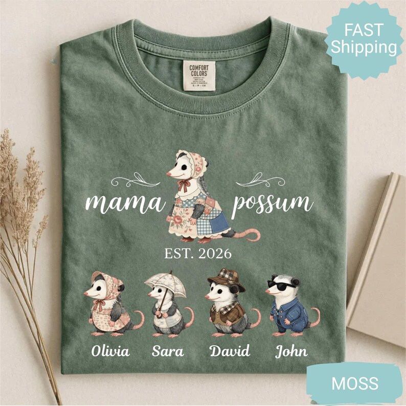 May include: Moss green t-shirt with a graphic of a possum family. The shirt features the text "mama possum" above an illustration of a possum wearing a bonnet and patchwork dress, with the text "EST. 2026". Below are illustrations of possum children.