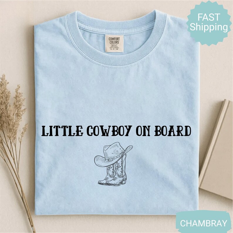 May include: Light blue t-shirt with the text "LITTLE COWBOY ON BOARD" in black, along with a graphic of a cowboy hat and boots. The shirt is made of a soft material and is likely for children.