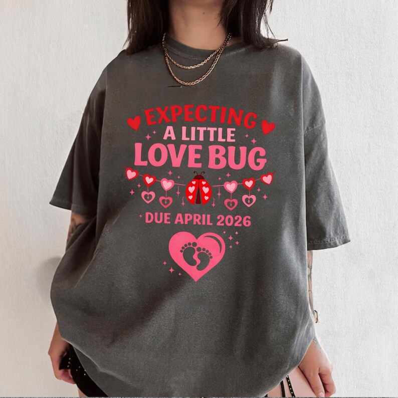 Op de afbeelding: Donkergrijs T-shirt met de roze tekst "EXPECTING A LITTLE LOVE BUG DUE APRIL 2026". Het ontwerp bevat harten, een lieveheersbeestje en babyvoetjes. Het shirt heeft een relaxte, oversized pasvorm.