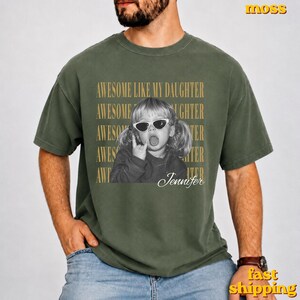 May include: A moss green short-sleeve t-shirt featuring a graphic of a child wearing sunglasses and making a hand gesture. The text AWESOME LIKE MY DAUGHTER is repeated in the background, and the name Jennifer is written in script below the child.