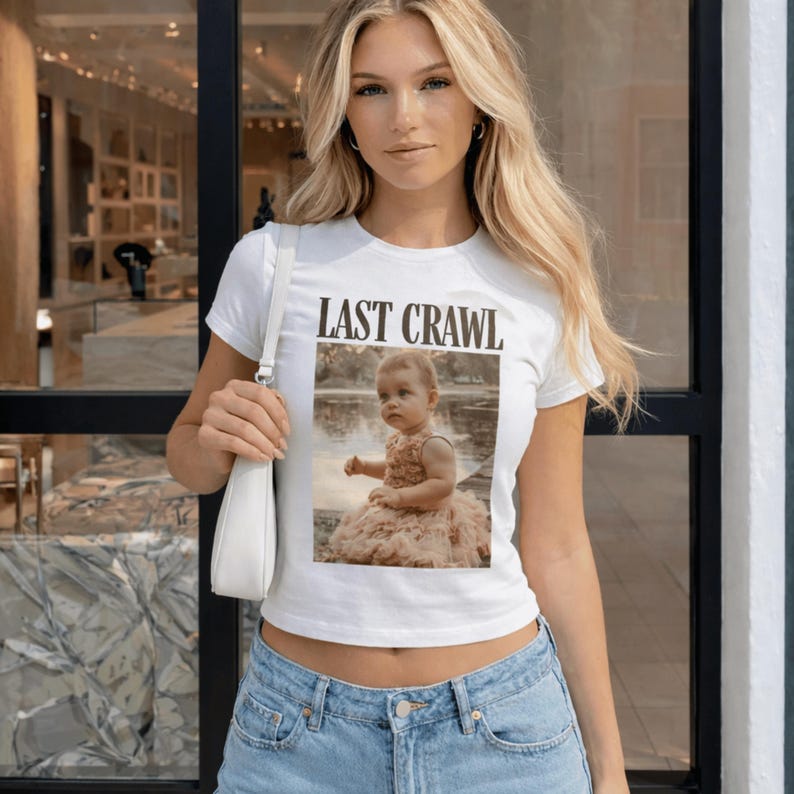May include: White t-shirt with the text "LAST CRAWL" above a photo of a baby in a dress. The shirt is short-sleeved and worn with blue jeans.