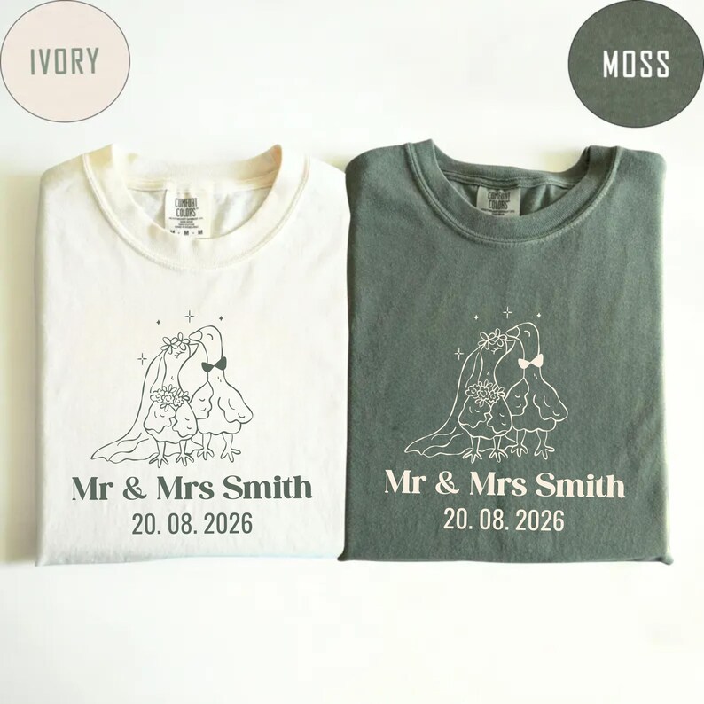 May include: Two t-shirts, one ivory and one moss green, with a wedding-themed design. Each shirt features a line drawing of two birds in wedding attire, the text "Mr & Mrs Smith 20.08.2026".