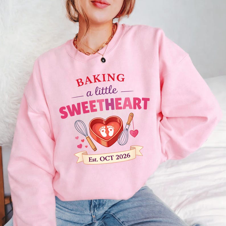 Op de afbeelding: Roze sweatshirt met de tekst "BAKING a little SWEETHEART". Het ontwerp bevat een hartvormige bakvorm met babyvoetjes, een garde en de datum "Est. OCT 2026".