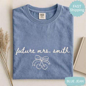 May include: A blue jean colored t-shirt with the text "future mrs. smith" in white script. Below the text is a white graphic of cherries with a bow. The shirt is folded and has a Comfort Colors tag.