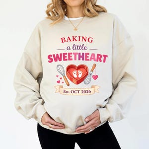 Op de afbeelding: Beige sweatshirt met de tekst "BAKING a little SWEETHEART" in rood en roze. Een hartvormige bakvorm met babyvoetjes is gecentreerd onder de tekst, geflankeerd door gardes en harten. De banner hieronder luidt "Est. OCT 2026".