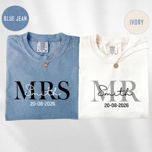 May include: Two t-shirts, one blue and one ivory, are displayed. The blue shirt reads "MRS Smith 20-08-2026" in black lettering. The ivory shirt reads "MR Smith 20-08-2026" in gray lettering. Each shirt has a gold necklace.