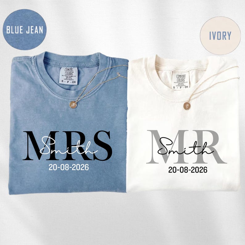 May include: Two t-shirts, one blue and one ivory, are displayed. The blue shirt reads "MRS Smith 20-08-2026" in black lettering. The ivory shirt reads "MR Smith 20-08-2026" in gray lettering. Each shirt has a gold necklace.
