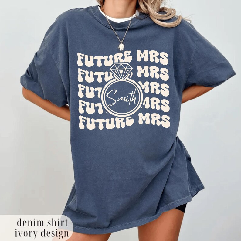 K&ouml;nnte beinhalten: Denimblaues T-Shirt mit dem vertikal wiederholten Schriftzug "FUTURE MRS" in Elfenbein. Eine Diamantring-Grafik mit dem Namen "Smith" ist zentriert. Das Shirt hat eine lockere Passform.