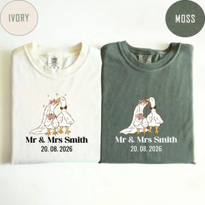 May include: Two t-shirts, ivory and moss green, with matching wedding-themed graphics. Each shirt features a cartoon goose couple with floral accents, a bow tie, and the text "Mr & Mrs Smith" and the date "20.08.2026".