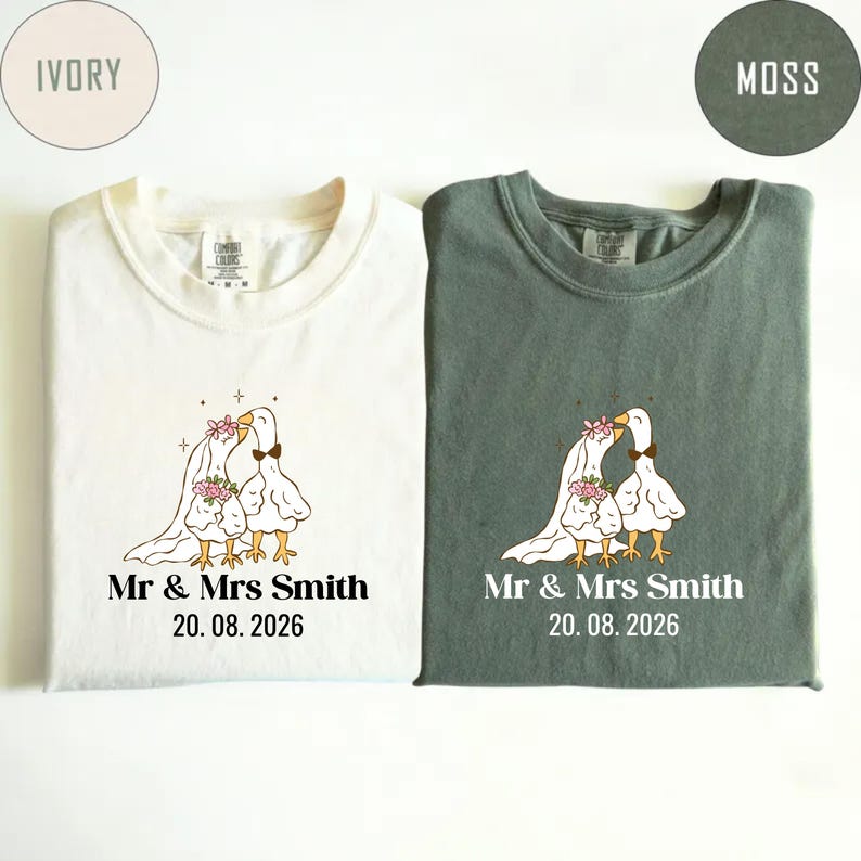 May include: Two t-shirts, ivory and moss green, with matching wedding-themed graphics. Each shirt features a cartoon goose couple with floral accents, a bow tie, and the text "Mr & Mrs Smith" and the date "20.08.2026".