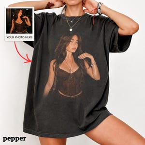 Puede incluir: Una camiseta gris carb&oacute;n de corte oversize con un gran estampado gr&aacute;fico centrado de una persona que lleva un top tipo cors&eacute; marr&oacute;n y un collar. La camiseta tiene mangas cortas y cuello redondo. Un peque&ntilde;o recuadro en la esquina superior izquierda muestra la imagen.