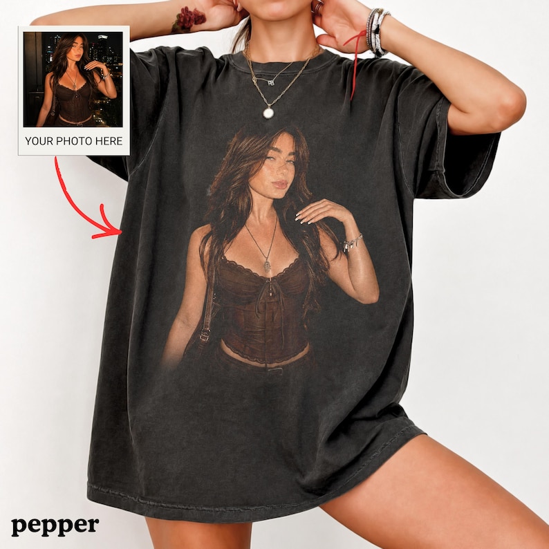 Puede incluir: Una camiseta gris carb&oacute;n de corte oversize con un gran estampado gr&aacute;fico centrado de una persona que lleva un top tipo cors&eacute; marr&oacute;n y un collar. La camiseta tiene mangas cortas y cuello redondo. Un peque&ntilde;o recuadro en la esquina superior izquierda muestra la imagen.