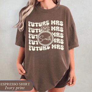 K&ouml;nnte beinhalten: Espressofarbenes T-Shirt mit elfenbeinfarbenem Aufdruck. Das Design zeigt die sich wiederholenden Worte "FUTURE MRS", mit einer Diamantring-Grafik und dem Namen "Smith" in der Mitte. Ein l&auml;ssiges Kurzarm-T-Shirt.