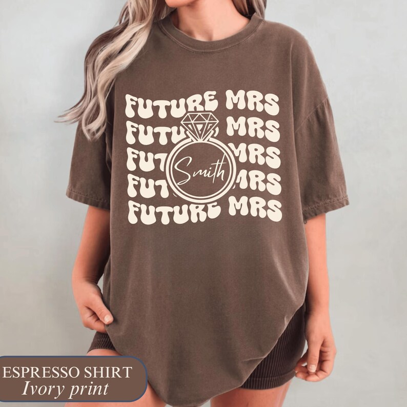 K&ouml;nnte beinhalten: Espressofarbenes T-Shirt mit elfenbeinfarbenem Aufdruck. Das Design zeigt die sich wiederholenden Worte "FUTURE MRS", mit einer Diamantring-Grafik und dem Namen "Smith" in der Mitte. Ein l&auml;ssiges Kurzarm-T-Shirt.