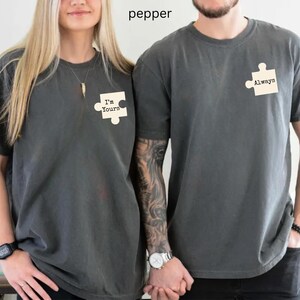 May include: Two matching pepper-colored t-shirts. One shirt has a puzzle piece with the text "I'm Yours", and the other has a puzzle piece with the text "Always". The shirts are short-sleeved and have a casual style.