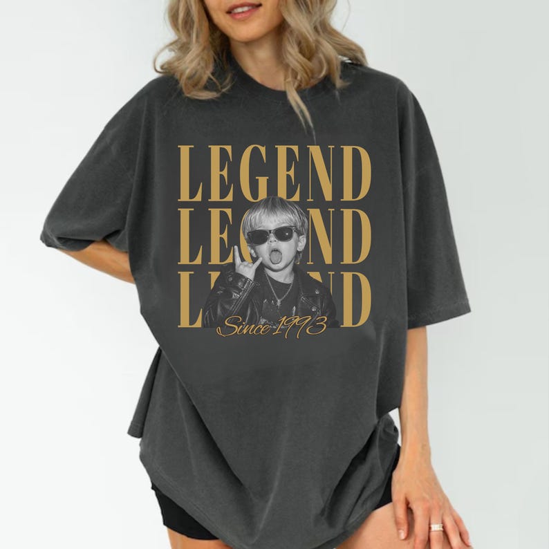 Puede incluir: Una camiseta gris carb&oacute;n de corte oversize con un estampado gr&aacute;fico de un ni&ntilde;o con gafas de sol y chaqueta de cuero. El texto "LEGEND" aparece en grandes letras doradas sobre y bajo la imagen, con el texto "Since 1993" en cursiva debajo.