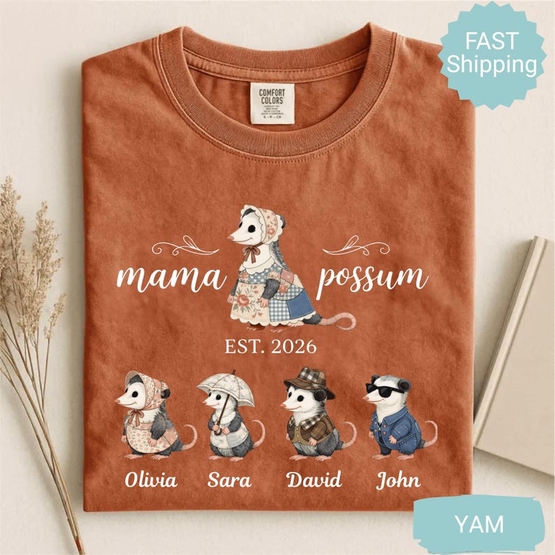 May include: Burnt orange t-shirt with a graphic of a possum family. The shirt features the text "mama possum" above an illustration of a possum wearing a bonnet and dress. Below are illustrations of possum children with names: Olivia, Sara, David, and John.