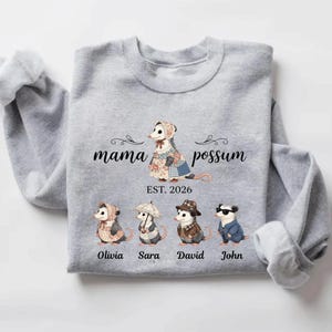 May include: A light gray sweatshirt featuring a "Mama Possum" design with a family of possums. The design includes the text "EST. 2026" and names: Olivia, Sara, David, and John. The possums are dressed in various outfits.