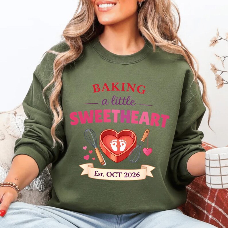 Op de afbeelding: Olijfgroene sweatshirt met de tekst "BAKING a little SWEETHEART" in roze en rood. Het ontwerp bevat een hartvormige doos met babyvoetjes, een garde en een spatel. De banner luidt "Est. OCT 2026".