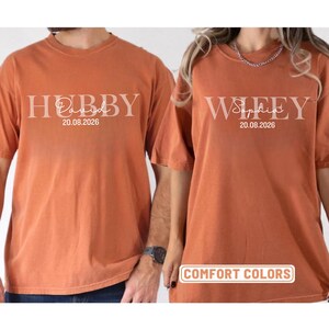 May include: Two burnt orange t-shirts with the words "Hubby" and "Wifey" printed in white, along with names and a date. The shirts are labeled "Comfort Colors".