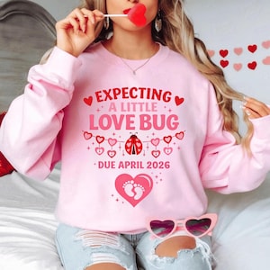 Op de afbeelding: Roze sweatshirt met de tekst "EXPECTING A LITTLE LOVE BUG DUE APRIL 2026". Het ontwerp bevat harten, een lieveheersbeestje en babyvoetjes. De sweatshirt heeft een lichtroze kleur.