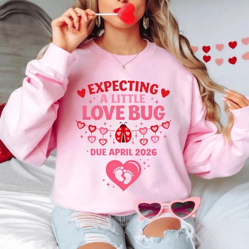 Op de afbeelding: Roze sweatshirt met de tekst "EXPECTING A LITTLE LOVE BUG DUE APRIL 2026". Het ontwerp bevat harten, een lieveheersbeestje en babyvoetjes. De sweatshirt heeft een lichtroze kleur.