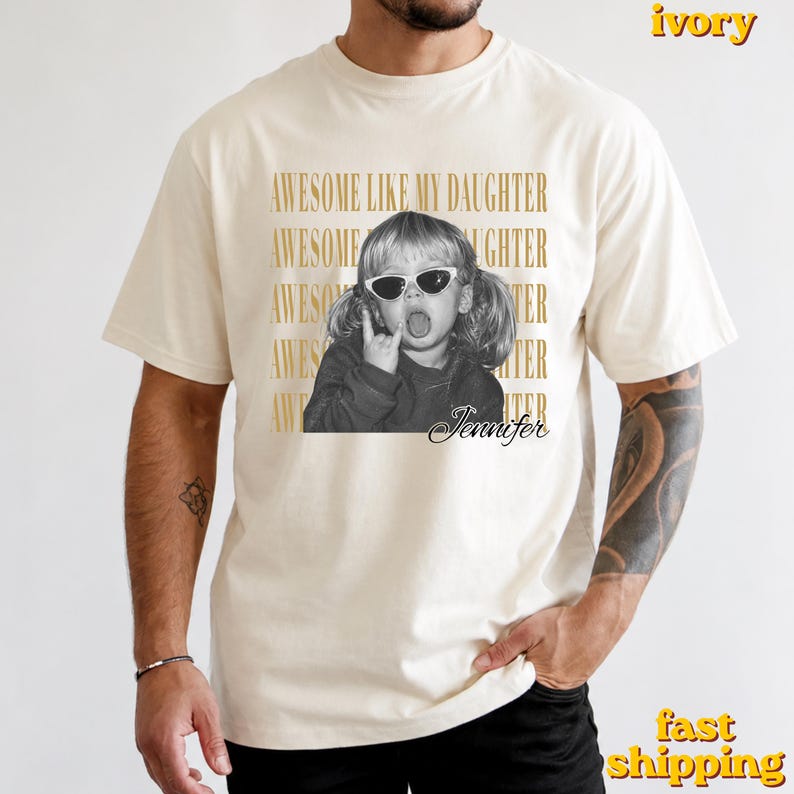 May include: An ivory short-sleeve t-shirt featuring a black and white graphic of a child wearing sunglasses and making a hand gesture. The text "AWESOME LIKE MY DAUGHTER" is repeated in gold behind the image, with the name "Jennifer" in script below.