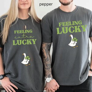 May include: Two dark grey t-shirts with the text "FEELING extra LUCKY" and "FEELING LUCKY" in green. Each shirt features a white goose holding a green four-leaf clover. The shirts are a casual style.