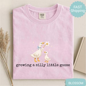 Może przedstawiać: Jasnor&oacute;żowa koszulka z ilustracją dw&oacute;ch gęsi, z kt&oacute;rych jedna trzyma stokrotkę. Pod spodem nadruk "growing a silly little goose". Okrągły dekolt i metka Comfort Colors.