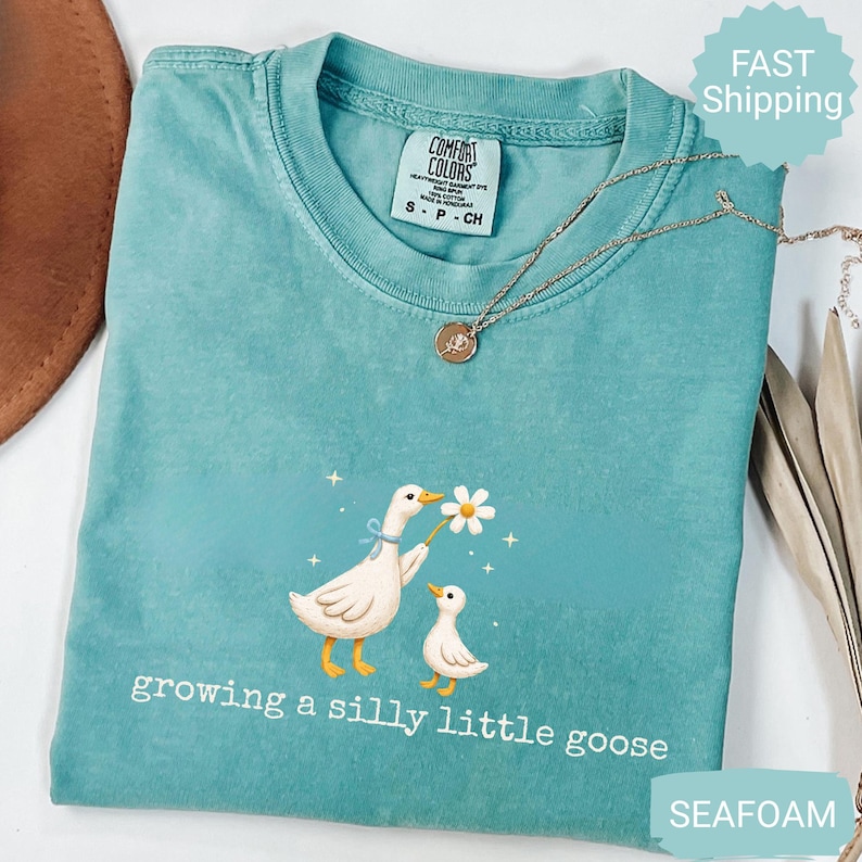 Może przedstawiać: Turkusowa koszulka Comfort Colors z ilustracją dw&oacute;ch gęsi, z kt&oacute;rych jedna trzyma stokrotkę. Poniżej znajduje się napis "growing a silly little goose". Złoty naszyjnik z wisiorkiem jest na koszulce.
