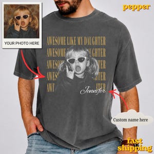 May include: A charcoal grey t-shirt featuring a black and white photograph of a child wearing sunglasses and making a hand gesture. The text AWESOME LIKE MY DAUGHTER is repeated in gold lettering behind the photo, with the name Jennifer in script.