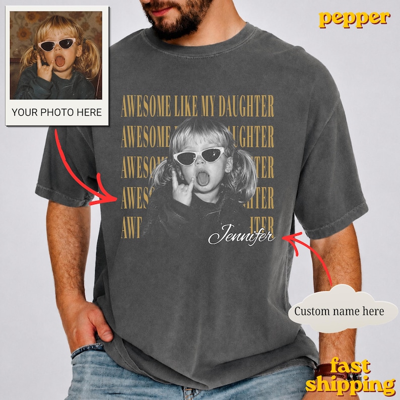 May include: A charcoal grey t-shirt featuring a black and white photograph of a child wearing sunglasses and making a hand gesture. The text AWESOME LIKE MY DAUGHTER is repeated in gold lettering behind the photo, with the name Jennifer in script.