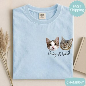 May include: Light blue t-shirt featuring two cat faces with the names "Daisy & Violet" in a script font. The shirt has a crew neck and a tag on the upper center. The shirt is made of a soft material.