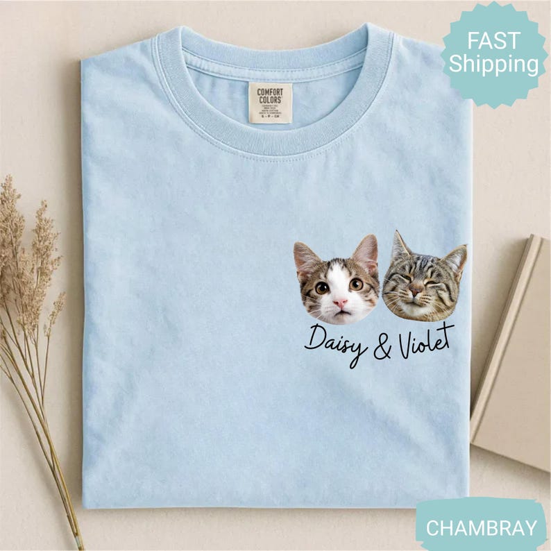 May include: Light blue t-shirt featuring two cat faces with the names "Daisy & Violet" in a script font. The shirt has a crew neck and a tag on the upper center. The shirt is made of a soft material.