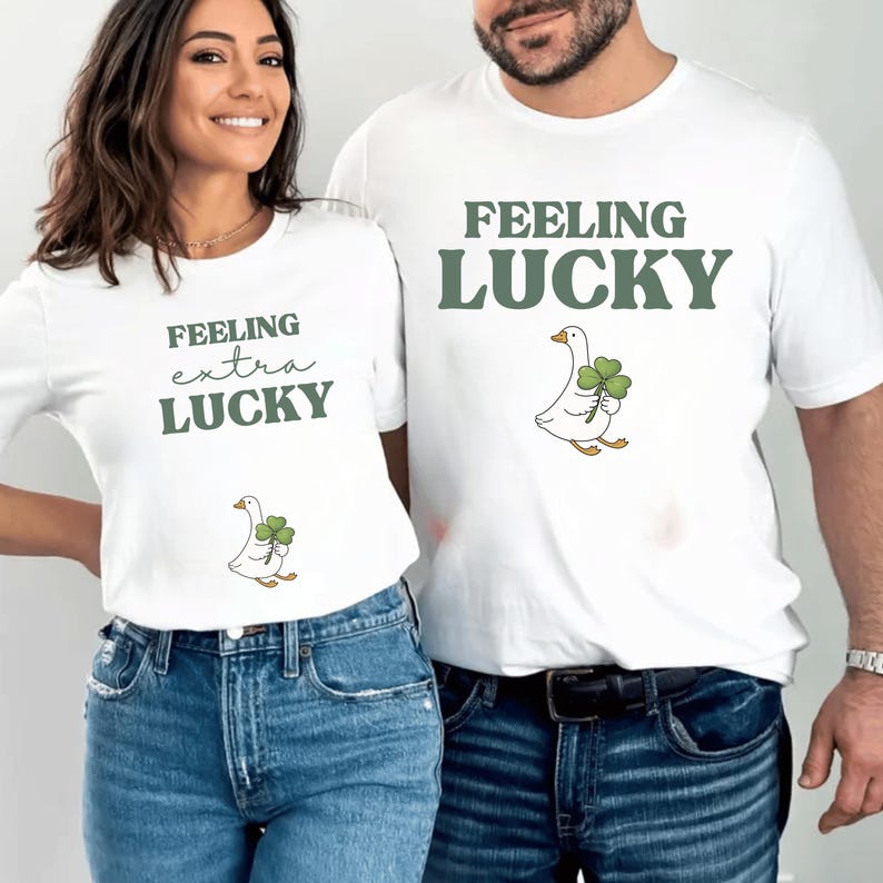 May include: Two white t-shirts with green text and a cartoon goose holding a four-leaf clover. One shirt says "Feeling extra lucky," the other "Feeling lucky." Both are worn with blue jeans.