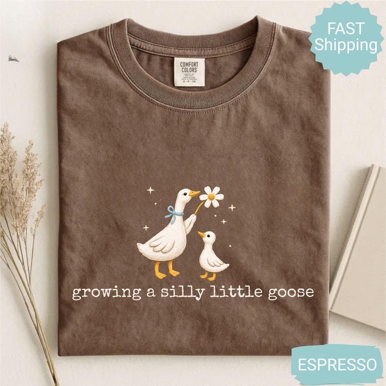 Może przedstawiać: Koszulka w kolorze espresso z fantazyjną ilustracją dw&oacute;ch gęsi. Jedna gęś trzyma stokrotkę, a pod spodem wydrukowano napis "growing a silly little goose". Wykonana z materiału Comfort Colors.