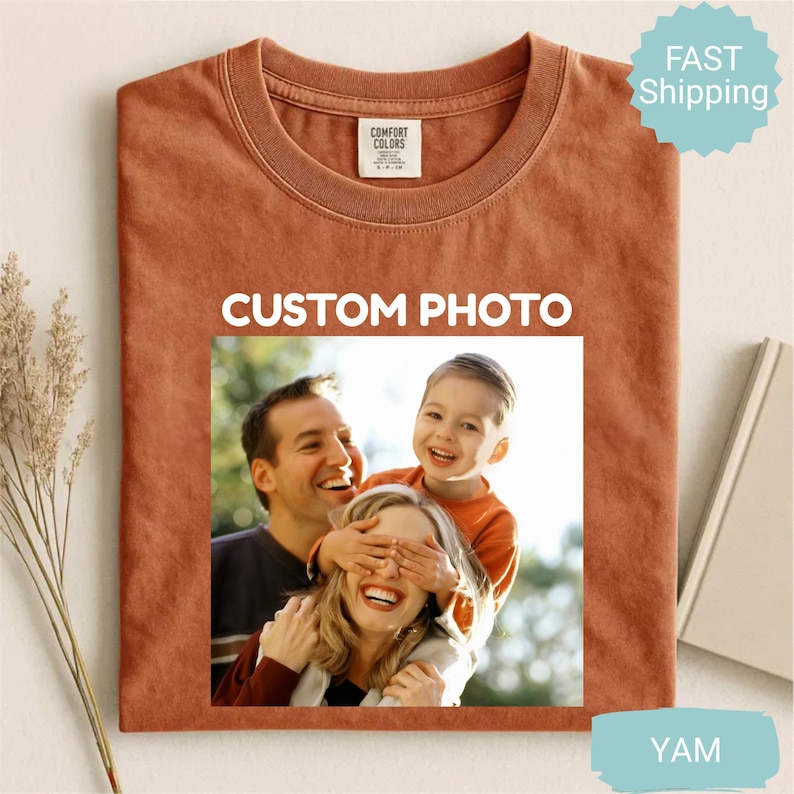 May include: A rust-colored t-shirt with a crew neck and a rectangular image of a family. The image on the shirt says "CUSTOM PHOTO" in white letters. The shirt has a tag that says "COMFORT COLORS".