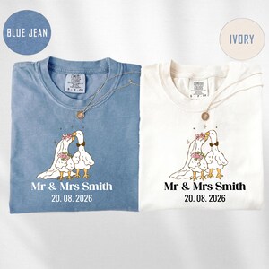 May include: Two t-shirts, one blue jean and one ivory, with a wedding-themed design. Each shirt features a cartoon goose couple, the text "Mr & Mrs Smith" and the date "20.08.2026". Both shirts have a crew neck and a necklace.