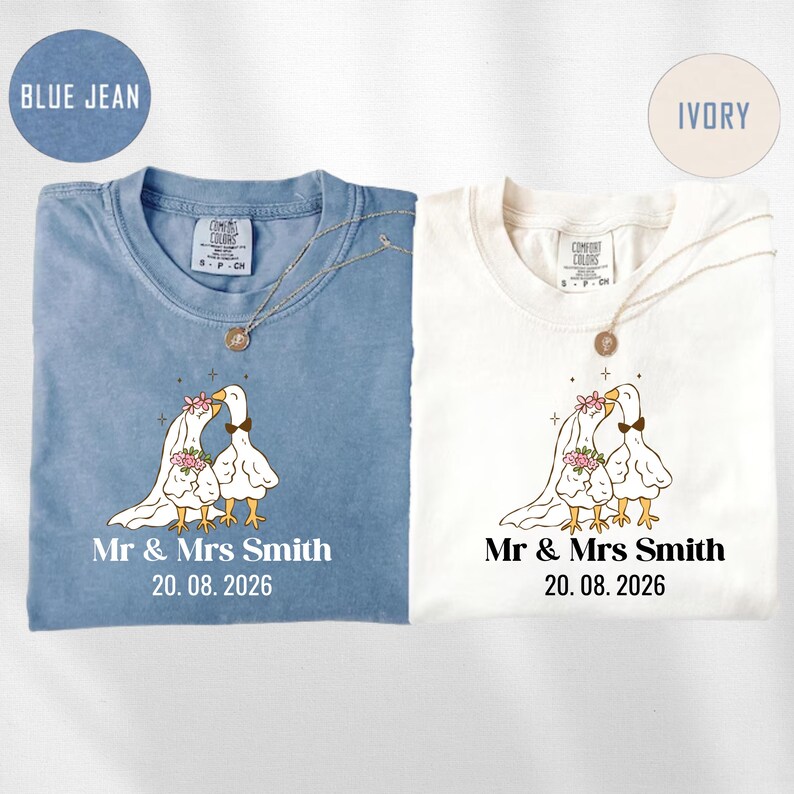 May include: Two t-shirts, one blue jean and one ivory, with a wedding-themed design. Each shirt features a cartoon goose couple, the text "Mr & Mrs Smith" and the date "20.08.2026". Both shirts have a crew neck and a necklace.