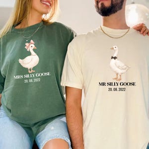 Peut inclure: Deux t-shirts assortis, un vert et un cr&egrave;me, avec une illustration d'oie. Le t-shirt vert dit "MRS SILLY GOOSE 20.08.2022" et le t-shirt cr&egrave;me dit "MR SILLY GOOSE 20.08.2022". L'oie sur le t-shirt vert a un n&oelig;ud rose, et l'oie sur le t-shirt cr&egrave;me a un n&oelig;ud papillon noir.