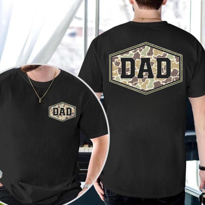 May include: Black t-shirt with a camouflage "DAD" design. The word "DAD" is in a bold, blocky font, set against a camouflage pattern within a hexagonal frame. The design is on the front and back of the shirt.