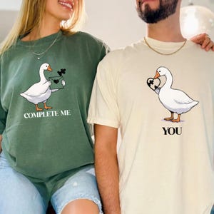 May include: Two matching t-shirts. One is olive green with a goose holding a puzzle piece and the text "COMPLETE ME". The other is cream-colored with a goose holding a heart-shaped puzzle piece and the text "YOU".