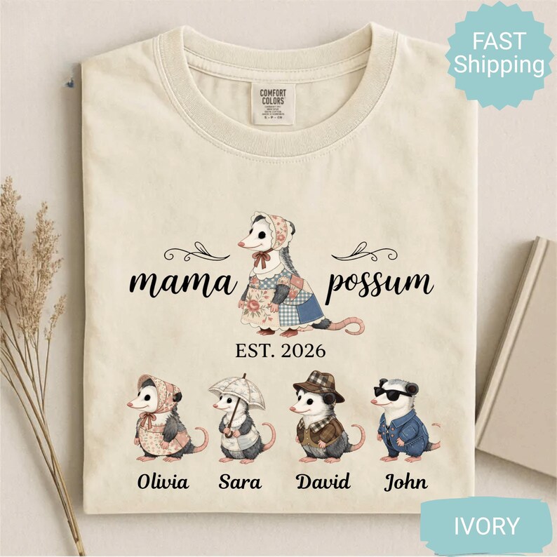 May include: Ivory-colored t-shirt featuring a graphic of a possum family. The design includes the words "mama possum" and the year "EST. 2026". Below are illustrations of four possums with names: Olivia, Sara, David, and John. The shirt has a crew neck.