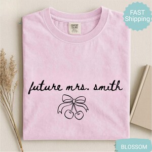 May include: A light pink t-shirt with the text "future mrs. smith" in black script, along with a bow and cherry graphic. The shirt is a crew neck style and is made of a soft material.