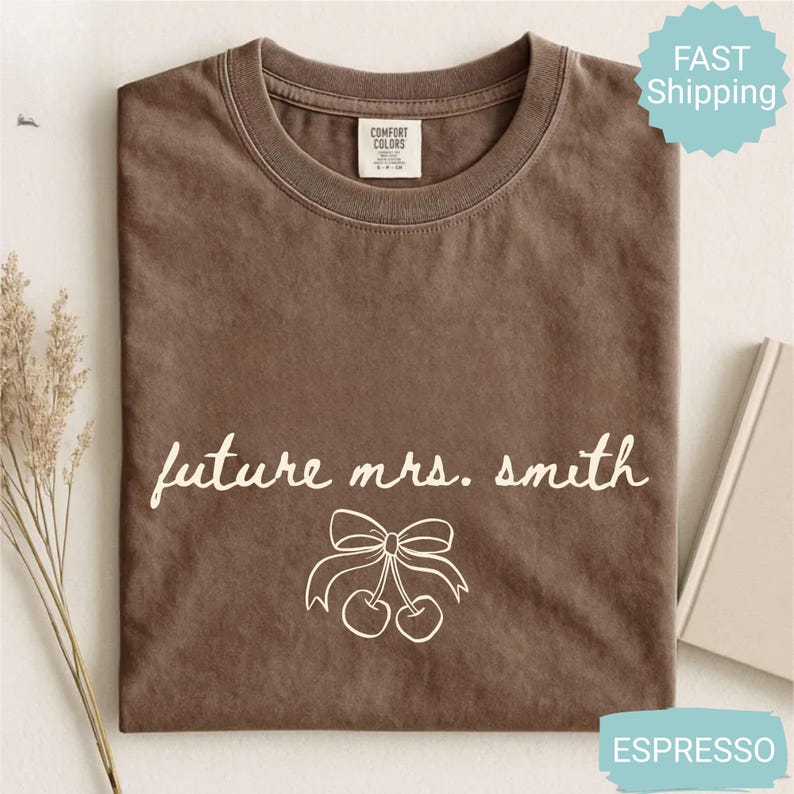 May include: Espresso-colored t-shirt with the text "future mrs. smith" in white script, along with a bow and cherry graphic. The shirt is folded and has a tag from Comfort Colors. The shirt is a crew neck.